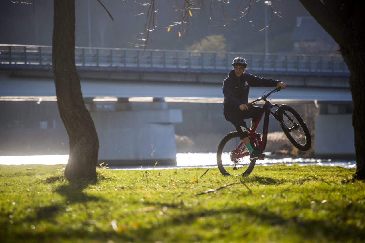 Rowerzysta jedzie na tylnym kole na rowerze full suspension. Świeci słońce a w tle widać rzekę